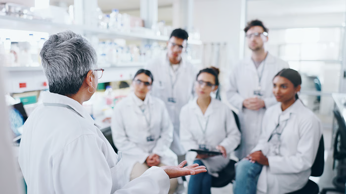 A group of five scientists, all wearing white laboratory coats, sitting on chairs listening intently to another scientist who is stood.