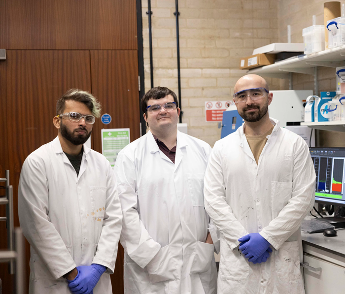 A group of three scientists from the Materials Group at the University of Birmingham, wearing goggles and laboratory coats, smiling at the camera.